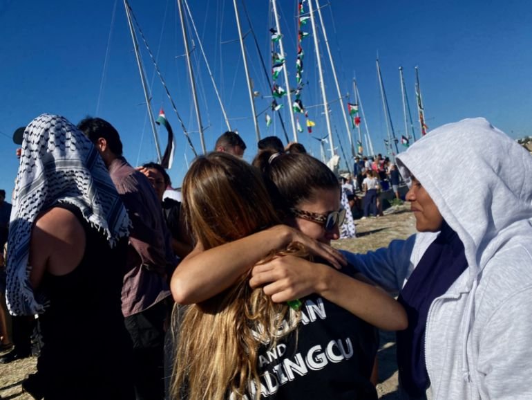 Female volunteers hugging their goodbyes with boats in the background, their masts full of fluttering Palestinian flags
