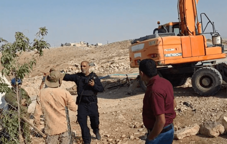 Israeli security officer shouts at Palestinians to keep them away from Yinon Levi who is in an excavator deliberately destroying the village's water and power supply lines