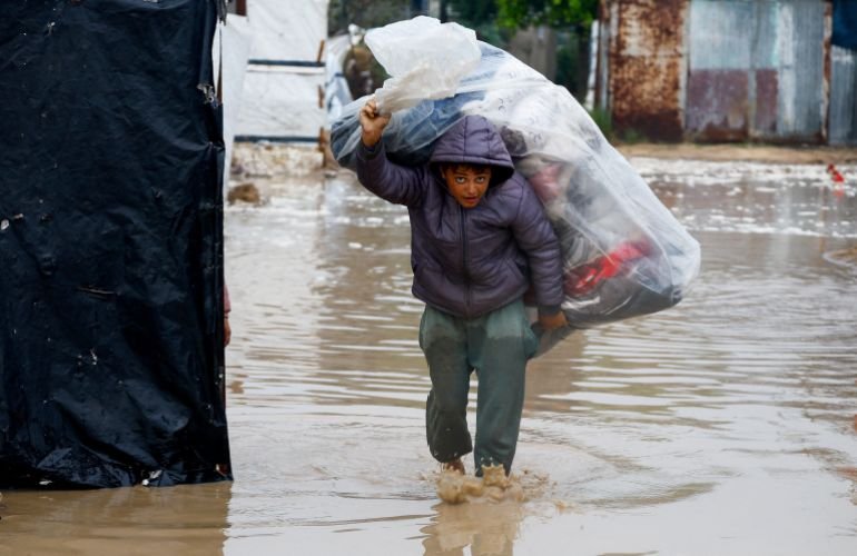 A displaced Palestinian boy carries belongings in a flooded tent camp on a rainy day in Nuseirat, central Gaza Strip, December 12, 2025. [Mahmoud Issa/Reuters]
