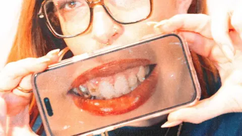 Billy-Jo Pierce A woman with ginger hair and glasses holds a phone up against the lower half of her face. The phone shows a picture of her smiling with her teeth as she shows off her tooth gems.