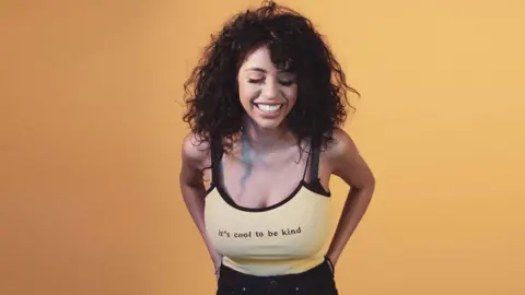 Ranvia  Ranvia smiling in front of an orange background, where she is stood wearing a strappy vest top which says 'it's cool to be kind.' The photo was taken before her breast reduction surgery