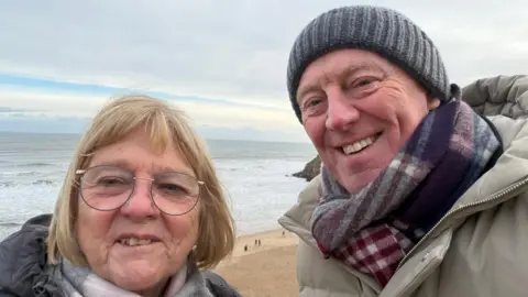 David Land David and Barbara, a couple in their sixties, on a beach in winter. David is wearing a grey woolly hat and plaid scarf. Barbara has blonde hair and glasses. Both are wearing winter coats.