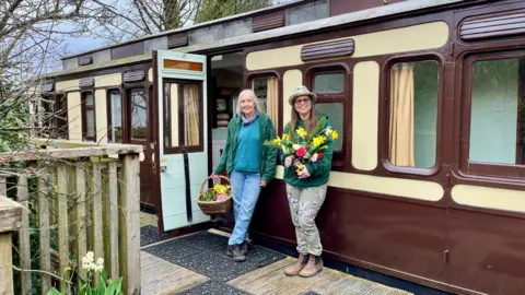 Lizzy Stroud Lizzy and Lil, two older women, standing outside an old train carriage which has been converted into self catering accommodation. Lizzy is holding a bunch of daffodils and Lil has a wicker basket of more flowers in her hand. 