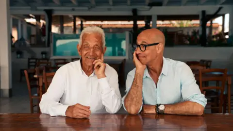 National Geographic/Matt Holyoak Tonino Bertoleoni and Stanley Tucci sitting at a table smiling 