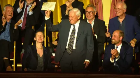 BBC Studios Sir David Attenborough holding up his letter from the King at the Royal Albert Hall, while seated next to Prince William