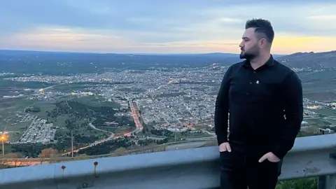 Kardo Jaf, also known as Kardo Ranya, stands at a roadside lookout beside a metal guardrail, wearing a fitted black long‑sleeved shirt and dark trousers, with hands resting in his pockets and body turned slightly to the right. The viewpoint overlooks a wide valley cityscape at dusk, with clusters of buildings, winding roads, and scattered streetlights stretching across rolling green hills. The sky is pale blue with soft clouds and fading daylight, and distant hills form the horizon in the background.