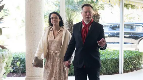 Getty Images Shivon Zilis holding hands with Elon Musk. Zilis is wearing a pale dress and jacket, and Musk is wearing a suit and a red scarf. Both are smiling.