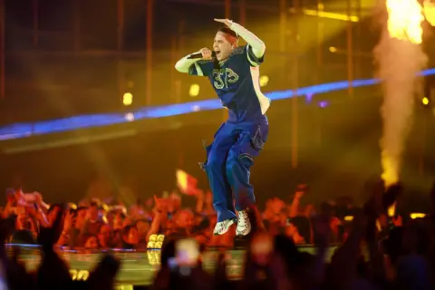 EPA Satoshi jumping in the air in a colourful tracksuit top, blue trousers and trainers 