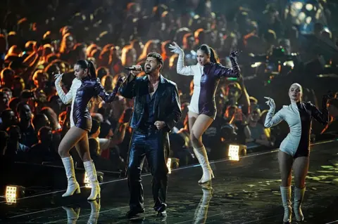 Getty Images Noam Bettan singing in a black outfit with three women dancing around him in black and white short outfits and white boots