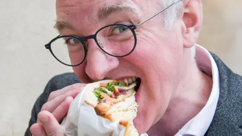 Emma Lynch/BBC The image shows a close-up of a person holding and eating a sandwich wrapped partly in white paper. The sandwich contains visible layers of bread, leafy greens, tomato, and slices of meat. The person is wearing round, dark-framed glasses and a textured jacket over a light-colored shirt. The background is softly blurred, drawing attention to the face and the sandwich in the foreground. The composition focuses tightly on the action of taking a bite, highlighting the textures of the food and the details of the glasses and clothing.