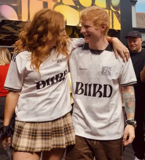 BIIRD 
Lisa Canny and Ed Sheeran stand close together outdoors with their arms around each other, both wearing matching white shirts with “BIIRD” printed on the front. Lisa wears a plaid skirt, while Ed has visible tattoos on one arm and is wearing brown trousers. A colorful sign or mural appears in the background, along with a few other people nearby.