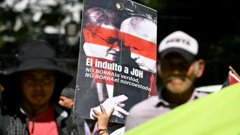 Orlando SIERRA / AFP via Getty Images) A woman holds a banner reading "The pardon for Juan Orlando Hernandez (JOH) does not erase the truth, does not erase the narco-state", during a protest  outside the US Embassy in Tegucigalpa on 4 December, 2025