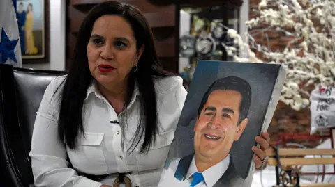 Orlando SIERRA / AFP via Getty Images Ana García Carías holds a painting of her husband during an interview with AFP in Tegucigalpa on 2 December 2. She is wearing a white shirt and red lipstick.  