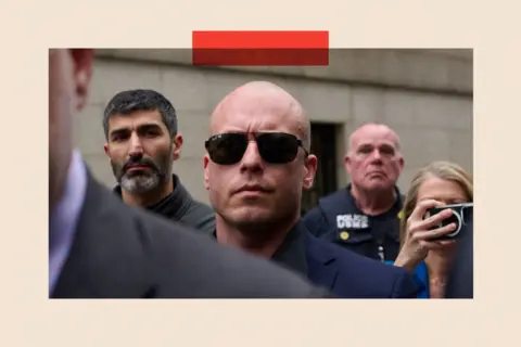 Getty Images A bald white man in sunglasses and a navy blue suit frowns. He is surrounded by people, including a police officer.