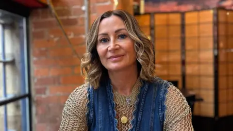 Sue Person seated at a restaurant table, arms resting on the surface, wearing a denim dress layered over a textured long-sleeve top, with a watch, rings, bracelets and a round pendant necklace. Sunglasses lie on the table to the left. Behind, a brick wall and decorative branches with pink blossoms and small warm fairy lights. A wooden room divider and a wall mural with a sunset scene appear in the background. Soft indoor lighting throughout.