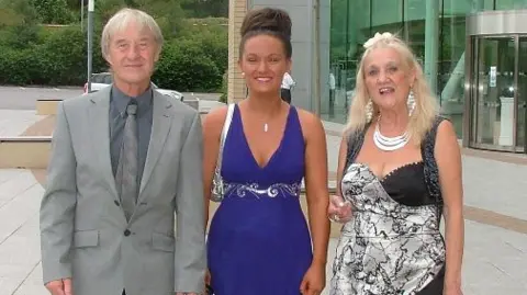 Michaela Allen Michaela with her grandmother at a wedding. She is stood in the middle of two older people. A man on her left, who is wearing a grey suit and tie, while Michaela wears a blue dress with a spiral pattern on. Her hair is tied up in a bun. Her grandmother is stood on her right and is wearing a grey and black dress. 