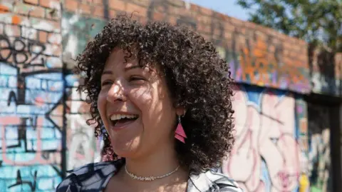 Handout Engy Elboreini smiles at the camera. She has curly dark-brown hair and pink earrings. She is standing in front of a wall spray-painted with graffiti.