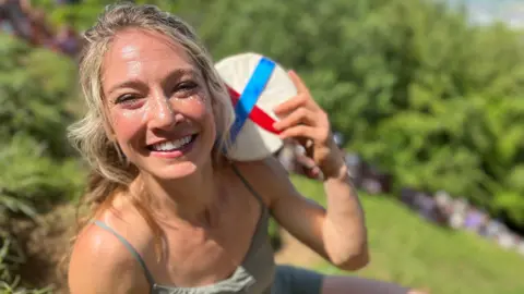 Ariel Dempsey holding a wheel of cheese after winning a cheese rolling race on Cooper's Hill. She has long blonde hair and is wearing a khaki green outfit. She is looking at the camera and smiling.