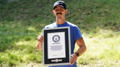 PA Media Cheese rolling contestant Chris Anderson with a Guinness World Records certificate. He was awarded for most wins in the Cooper's Hill cheese rolling and wake race. Chris is wearing a blue cap and t-shirt and he is holding the certificate in front of his chest. He is smiling. 