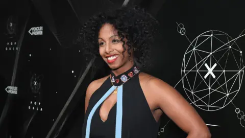 Getty Images Luam wears a black halter neck dress with a blue ribbon on the front and flowers on the collar. She wears red lipstick and has her black curly hair in a short style 