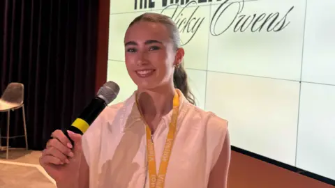 Vicky Owens Vicky wears a white shirt with a yellow lanyard and holds a microphone as she smiles at the camera. She has long brown hair which is up in a ponytail. In the background is a big white screen and events area