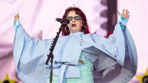 Getty Images CMAT performs on Radio 1's Big Weekend stage against a pink backdrop with yellow stars. She wears a light blue floral two-piece outfit with a blue striped cape and has shoulder length red hair. She wears silver hoops and sunglasses and holds up her hands to the crowd