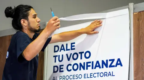 AFP via Getty Images An election worker with dark hair tied in the back, a beard and a large earring is seen in profile as he sticks a poster at an election centre