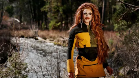 Evan Benally Atwood Performer Wyn Wylie, in drag as Pattie Gonia, wearing a yellow weatherproof dress with heavy face make up and long ginger hair. 