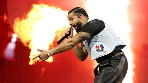 Getty Images Drake performs on stage as a plume of flame erupts behind him, framing the star as he raps into a microphone and points into the crowd with his free hand. Wearing a white vest with a cartoon image of a bird printed on it, he looks intense.