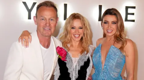 PA Media Kylie Minogue (centre) pictured with her arms around her sister Dannii and actor Jason Donavan