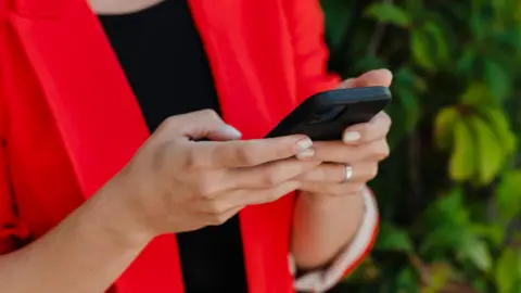 Getty Images Close up of a woman's hands holding a black smartphone. She is wearing a bright red jacket and a silver ring.