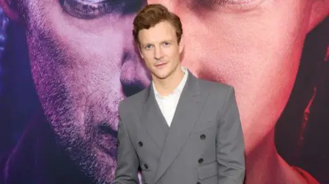 WireImage Patrick Gibson in a grey suit on a red carpet.