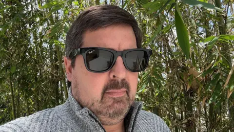 Mark Smith A man standing outside in a grey sweater and wearing Meta's Ray-Bans with a wall of green bamboo behind him. 
