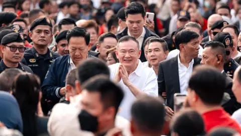 Thai News Pix Thaksin, smiling and holding his hands in a prayer pose, stands among a sea of supporters