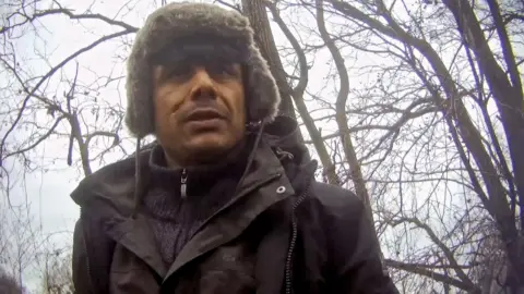 Zia is wearing a fur-lined winter hat and dark jacket standing outdoors among leafless trees on an overcast day.