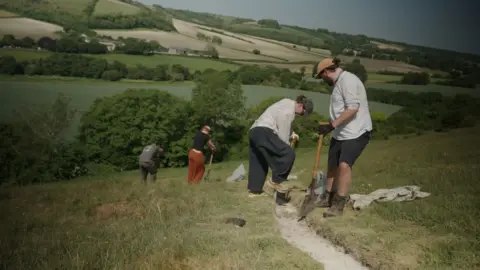 BBC/ Kevin Church A person with a ponytail wearing a hat, a long shirt and long trousers is digging into the white chalk with one leg on top of a spade. A man wearing a hat, a t-shirt and shorts also carrying a spade is looking on next to the person. A couple of other volunteers are seen in the distance. Beyond them, you can see a line of trees and more green fields in the background.   