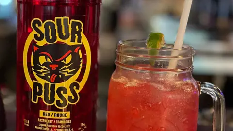 Courtesy of Phillips Distilling Company An image showing a close-up of a red Sour Puss bottle, with a black and yellow logo that features a drawing of a cat sticking its tongue out. Next to the bottle is a red cocktail in a mason jar glass with a plastic straw and a lime.