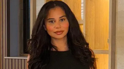 Rochelle Lewis A woman wearing a black outfit stands indoors against a softly lit background with shelves and warm lighting.
