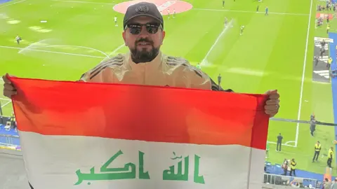 Abdulla Adnan A man wearing a dark baseball cap and sunglasses holds up an Iraqi flag in a stadium. A football pitch can been seen behind him. 