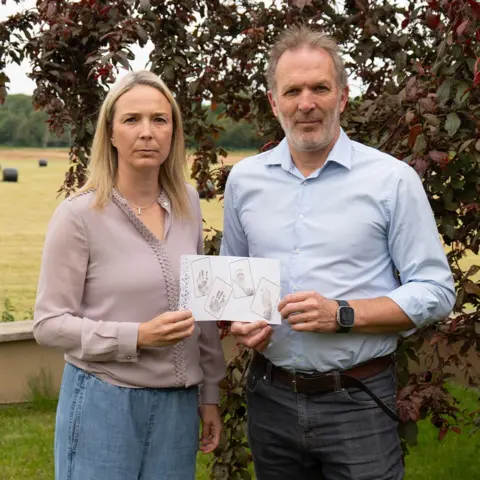 BBC/Chris Waring Sarah and Jack Hawkins stand side by side, holding their daughter's handprints and footprints