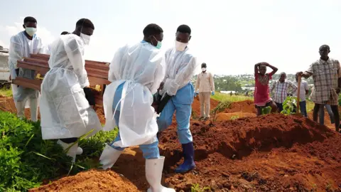 Amensisa Ifa / BBC Volunteers in blue scrubs, rubber boots and white gowns carry a coffin towards a dug grave. A few people, including one holding a spade, watch on from the side