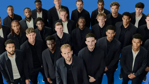 Marks & Spencer England men's squad wearing dark jackets and trousers, standing with their arms folded in front of them 