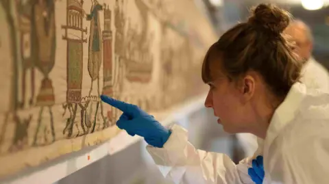 French Ministry of Culture A woman in a white hazmat suit and blue gloves puts a finger to the tapestry.