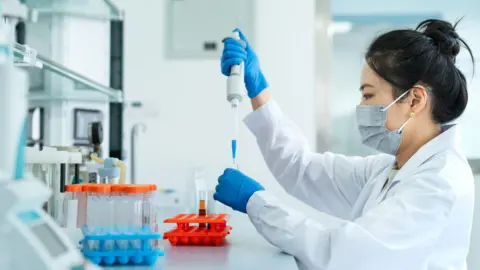 Getty Images A laboratory setting with a person in a white lab coat working at a bench. The person is wearing protective gloves and a face mask while carefully using a pipette to transfer liquid into a small test tube. Several racks of test tubes with coloured liquids are arranged neatly on the work surface in the foreground. Scientific equipment and shelves are visible in the background, suggesting a clean and organised workspace