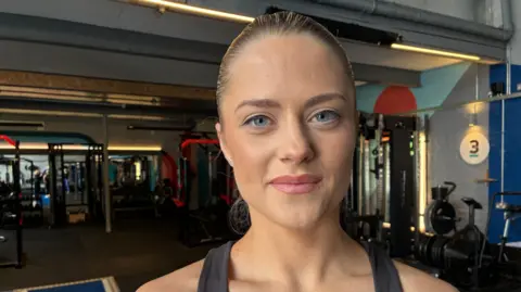 BBC/Lesley Hitchen A dark haired woman with her hair sleeked back behind her head wears workout clothes and stands in a gym. She is looking directly at the camera and smiling. 
