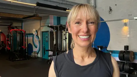 BBC/Lesley Hitchen Bev Wilson is in a gym, wearing workout clothes. She is smiling at the camera and has blonde hair in a ponytail. 
