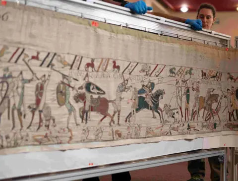 French Ministry of Culture The tapestry attached to a concertinaed frame with specialists wearing blue gloves behind it.