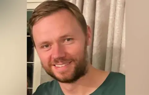 Ministry of Defence/PA Wire Lt Cdr Chris Gayson. He his pictured wearing a green top and is looking in the direction of the camera. He is smiling. He is standing in front of a curtain. 