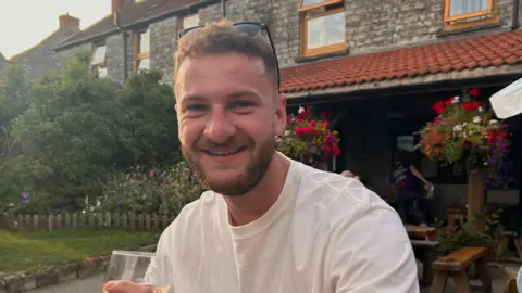 Ministry of Defence/PA Wire Petty Officer Owen Green. He is pictured in a white top is sitting at a bench in the outdoors. A stone building is behind him. He is holding a wine glass and is smiling at the camera.