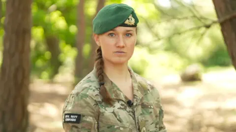 Lt Lily-Mae Fisher. She is pictured in military uniform and wearing a beret is standing outdoors under trees. She is looking in the direction of the camera. The background behind her is blurred. 